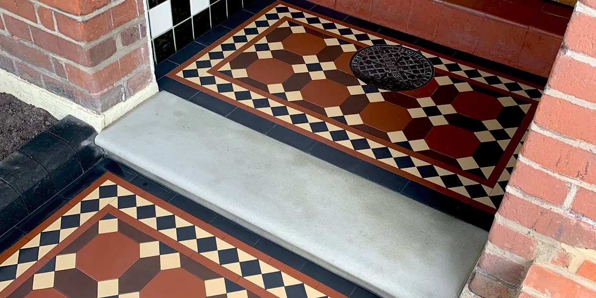 A porch with stone step tread and glazed tiles to the side. The Edwardian-style tile pattern continues from the path to the porch step. An antique coal-hole cover sits in the centre of the porch step.