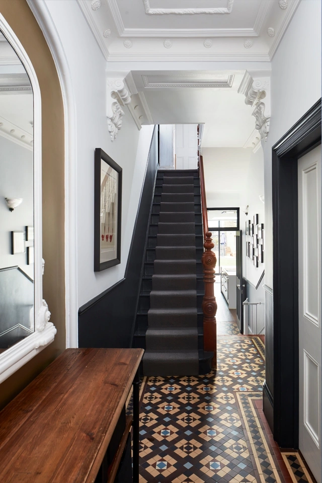 A brown and yellow toned Victorian-style mosaic floor pattern in a high-ceilinged hallway. To the left a mirror is fitted in an alcove with a wooden table beneath.A black painted staircase, with black panelling rises. To the rear part of a kitchen and patio doors are seen. On the ceiling ornate cornices.