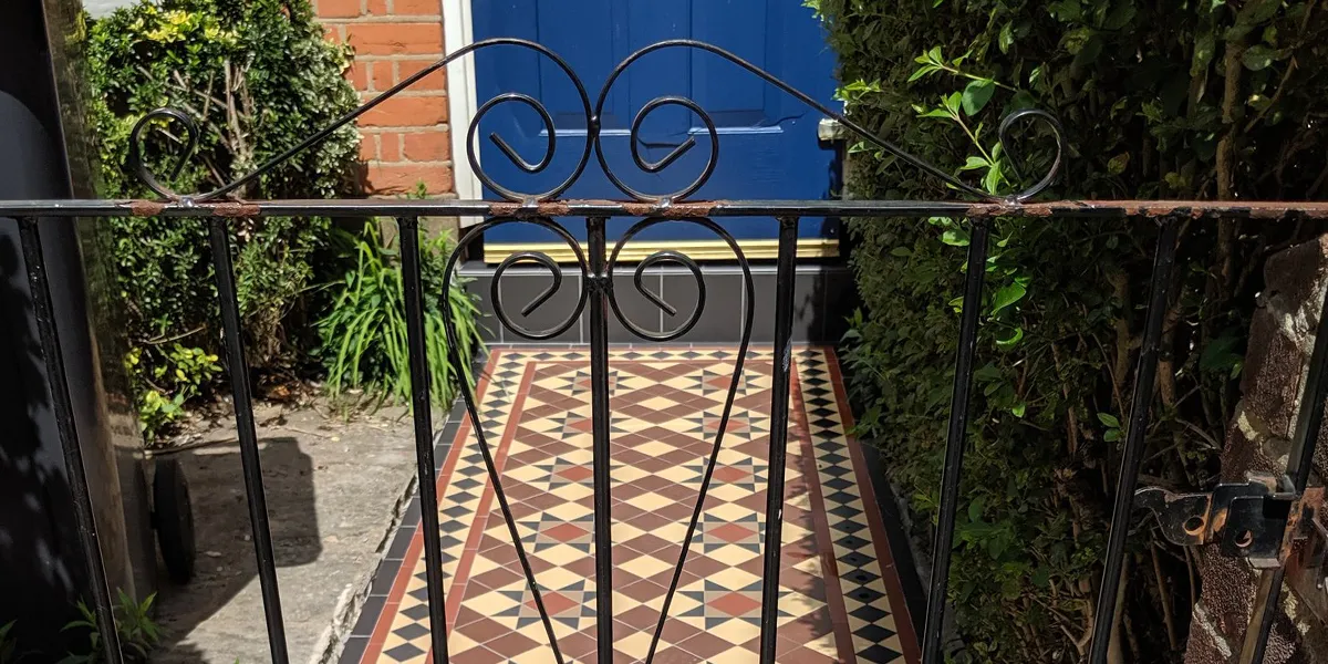 Viewed through a closed wrought-iron gate, an Edwardian style path leads to a deep blue front door. To the right of the path a tall hedge, and to the left more green foliage and a black bin cast a shadow on the concrete floor.