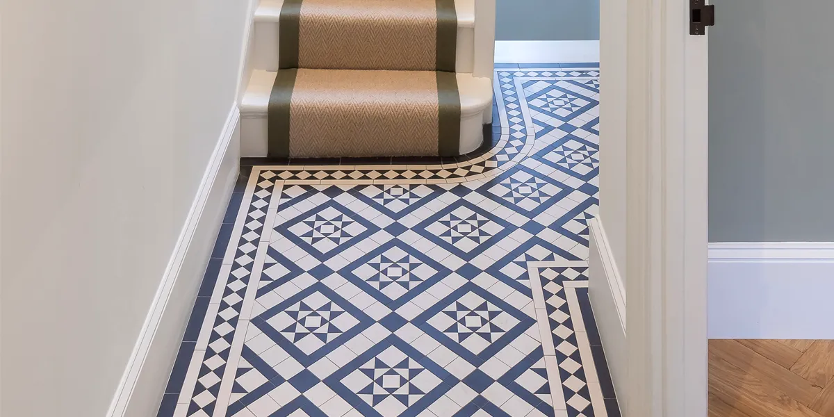 A decorative black and white mosaic floor tile pattern in a small hallway. The border design curves around the base of the staircase. The woodwork is painted gloss white, and there is a beige and green stair runner.