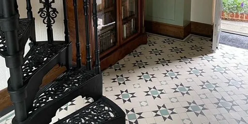 A simple star-motif Georgian mosaic tile pattern in black, white and green. To the left, a decorative wrought-iron spiral staircase rises. Behind this we see a dark wood bookcase with glass doors. To the right, the front door is open revealing some flowering shrubs.