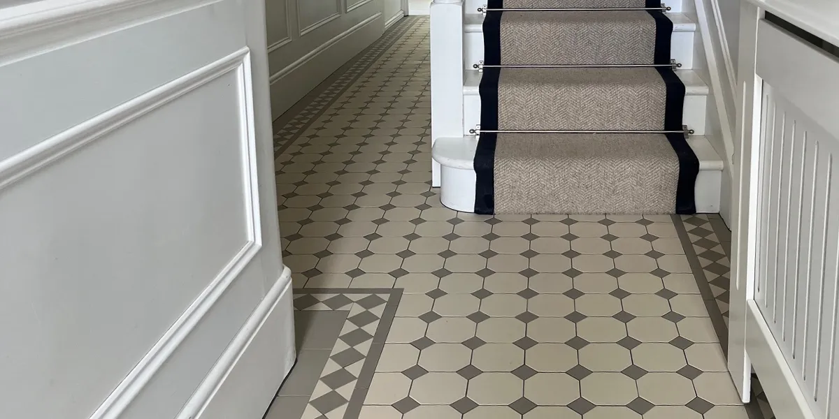 A narrow hallway with grey and white, octagon and dot tiled floor. The walls have white painted panelling, and the right a radiator is hidden behind a white-painted wooden cover. The staircase has a grey stair-runner.