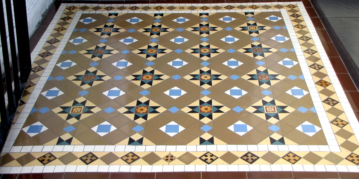 A Victorian mosaic path with antique encaustic tiles. The intricate brown, yellow and blue design is framed by a traditional border motif. To the left, a black metal gate is open.