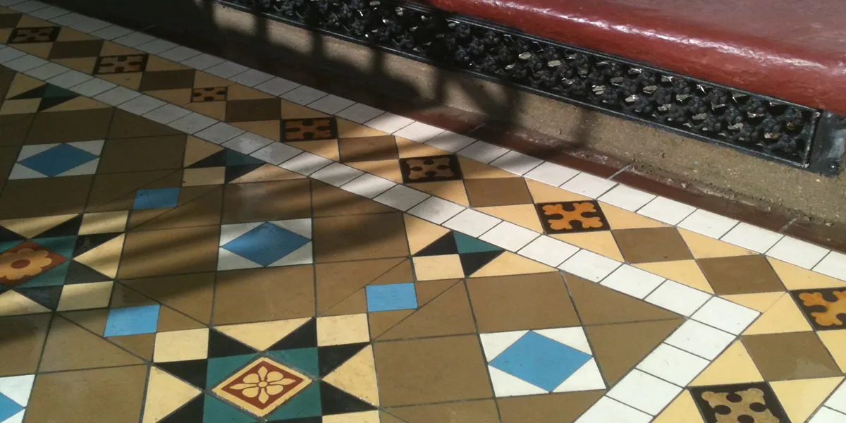 Close view of a mosaic tiled path which features several encaustic tiles. A red painted step is seen behind the path, with a daisy grate ventilation grille fitted below.