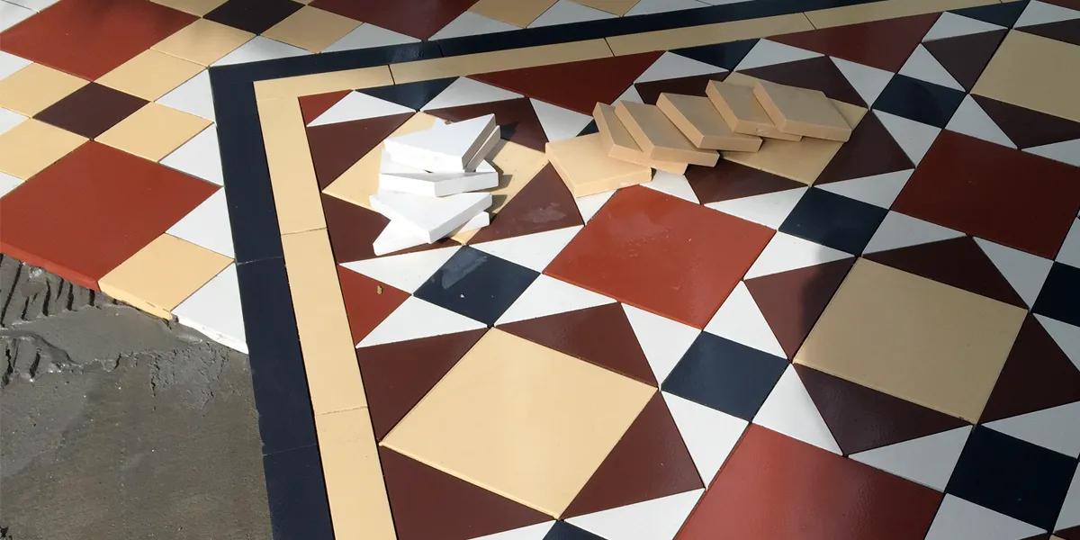 Close up view of some Edwardian style mosaic tiles in black, white, red, brown and yellow colours. The pattern is half installed, and the pattern uses squares, triangle and rectangles. There are some white triangles and yellow squares sitting on top of the installed tiles.