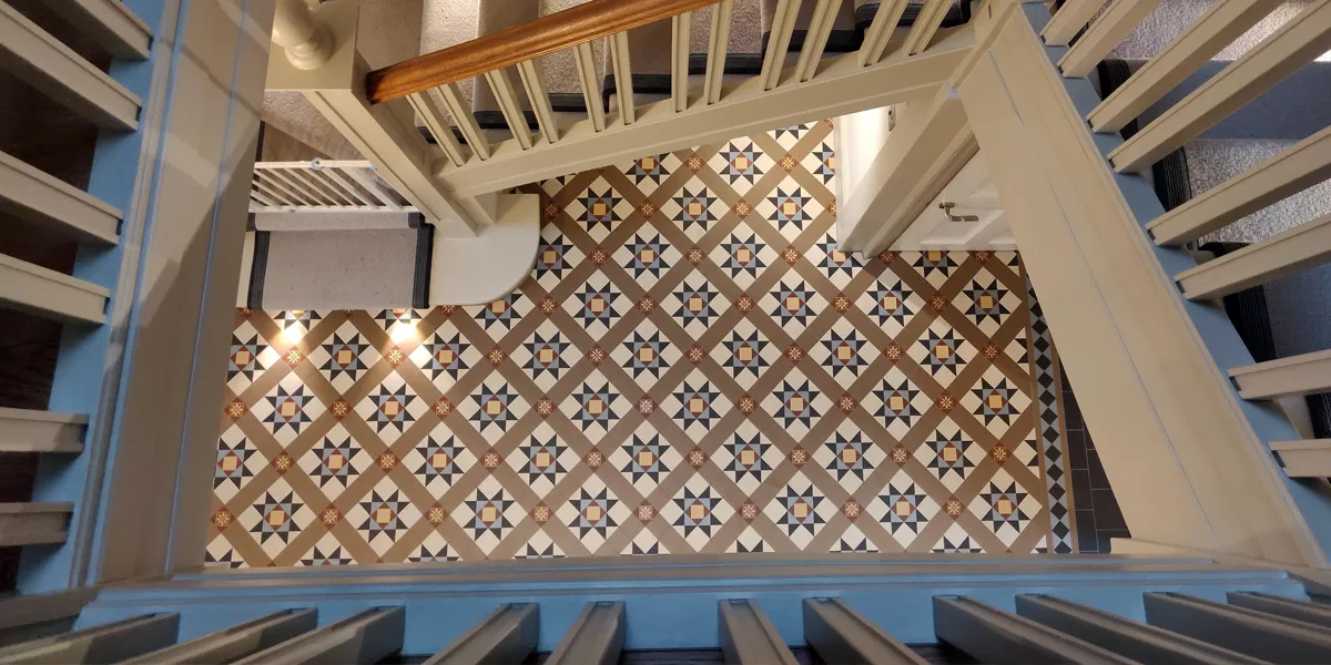View from the second floor looking directly down a staircase to a Victorian-style mosaic floor in the hallway below.