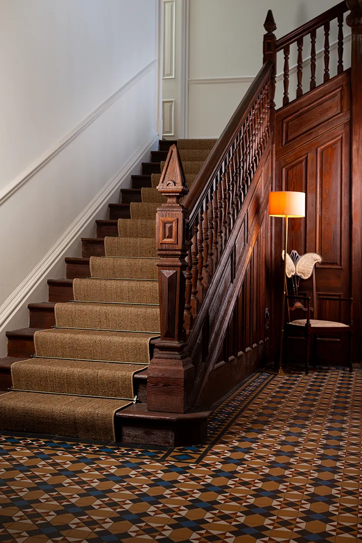 A large dark-wood staircase with ornate balstrade rises within a large high-ceilinged hallway. The staircase has a hesian runner. A lamp and art-nouveau style chair sit at its base. The floor of the hallway is highly decorative Victorian style mosaic in yellow and blue tones. A repeating star motif sits within a trellis pattern.