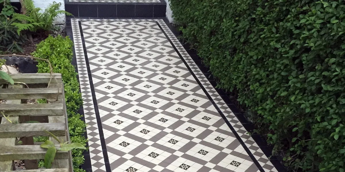A traditional Victorian trellis patterned mosaic pathway, in grey, white and black tiles. The design includes a black and white encaustic tile with a floral motif. The path design is framed to the left and right with a border pattern of diamond set grey squares and white triangles. To the right of the path a large green hedge. To the left, the garden is a bit overgrown, and an old wooden frame is seen to the front left-hand side.
