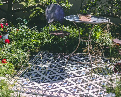 Victorian tiles used as garden patio feature. The Bexley design in black, white, blue and yellow coloured geometric tiles. Metal table and garden chairs sit on tiled patio surrounded by wildflower borders.