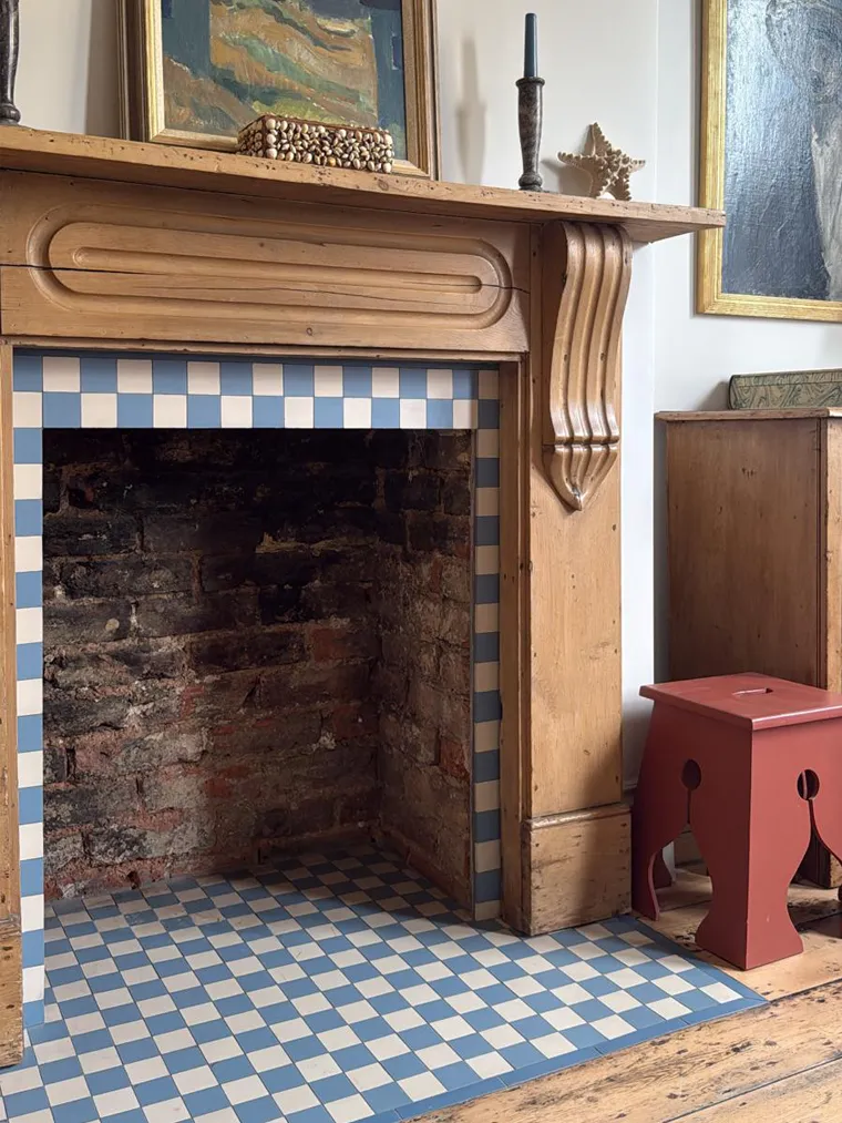 Blue and white chequered tiles decorate a fireplace hearth and surround. Internally, the fireplace is exposed brickwork. It has a waxed, oak-wood mantle and legs. To the right of the fireplace there is a wooded stool painted in a light burgundy shade.