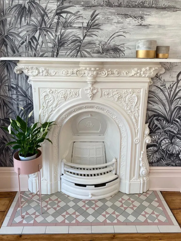 A higly decorative, antique cast-iron fireplace is painted white. In front there is a hearth with Victorian-type mosaic tiles but in pastel shades of white, pink and pale grey. To the left-hand side of the hearth, a plant sits in a light pink planter, with spindle legs. The wallpaper behind the fireplace is a richly illustrated black and white scene of bamboo and other plants.